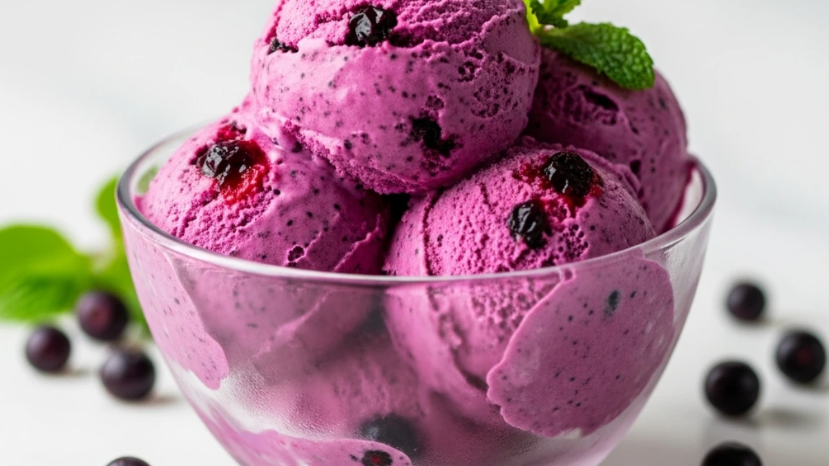 Bowl of vibrant purple huckleberry ice cream with fresh mint garnish and scattered huckleberries on white marble.